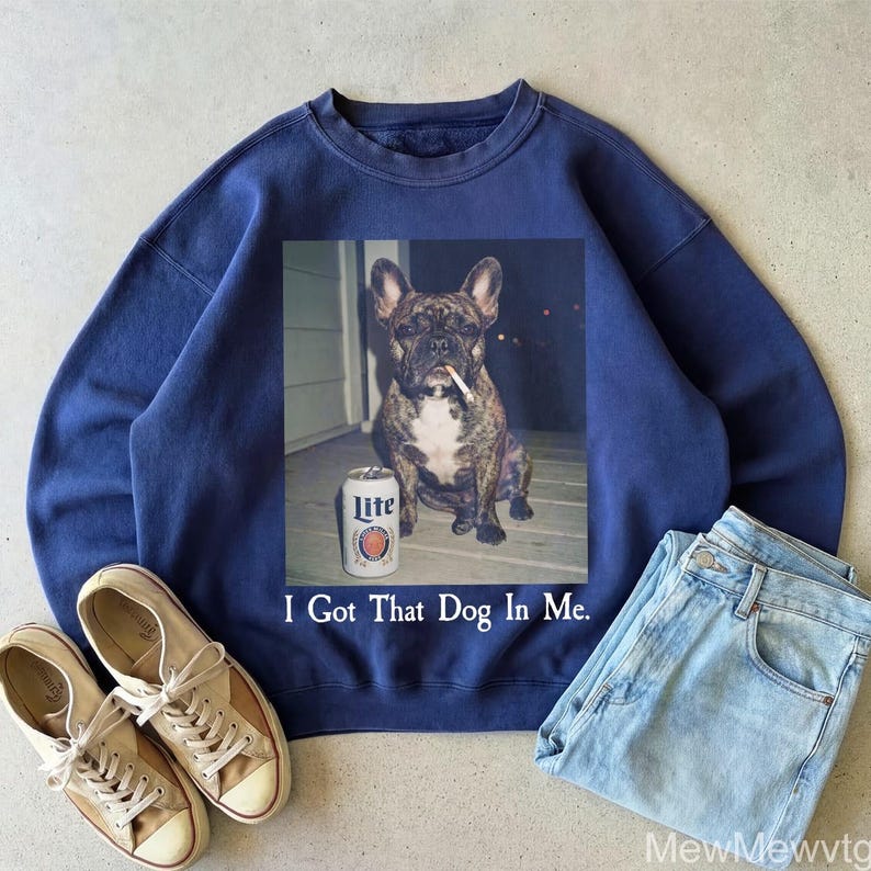 May include: Navy blue sweatshirt featuring a graphic of a brindle French Bulldog with a cigarette and a can of Lite beer. The text "I Got That Dog In Me" is printed below the graphic. The sweatshirt is paired with light wash jeans and canvas sneakers.