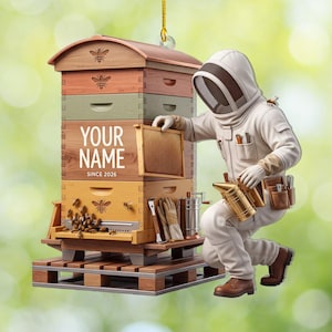 May include: A hanging ornament featuring a multi-tiered wooden beehive with the text YOUR NAME SINCE 2026 and a beekeeper in a white protective suit kneeling beside it. The beekeeper holds a honeycomb frame and a metal smoker, with bees visible.