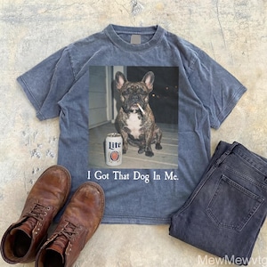 May include: A blue t-shirt with a graphic of a French bulldog with a cigarette in its mouth and a can of Lite beer. The text "I Got That Dog In Me" is below the graphic. Brown leather boots and dark blue jeans are also in the image.