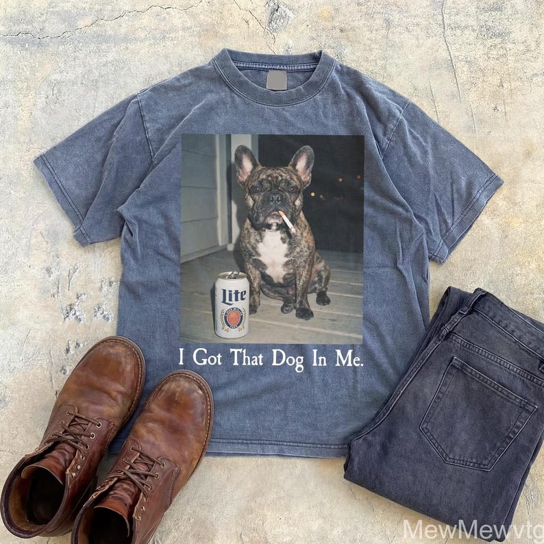 May include: A blue t-shirt with a graphic of a French bulldog with a cigarette in its mouth and a can of Lite beer. The text "I Got That Dog In Me" is below the graphic. Brown leather boots and dark blue jeans are also in the image.