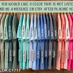 May include: A display of Comfort Color C1717 t-shirts in various colors, including white, mustard, blue spruce, and black, hanging on wooden hangers. The image includes text that reads, "If you would like a color that is not listed, please send us a message on Etsy after placing your order."