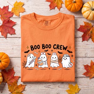 May include: An orange t-shirt featuring a graphic of four cartoon ghosts dressed as medical professionals with the text "BOO BOO CREW" above them. The ghosts are depicted with medical items including a thermometer, a bottle, and a first aid kit.