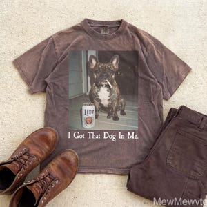 May include: A brown t-shirt with a graphic of a dog smoking a cigarette and a can of Lite beer. The text "I Got That Dog In Me" is below the graphic. Brown work boots and brown pants are also in the image.