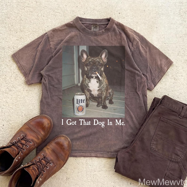 May include: A brown t-shirt with a graphic of a dog smoking a cigarette and a can of Lite beer. The text "I Got That Dog In Me" is below the graphic. Brown work boots and brown pants are also in the image.