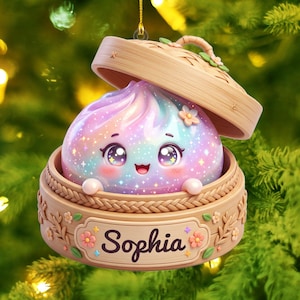 Könnte beinhalten: Ein hängendes Ornament mit einem pastellfarbenen, irisierenden Knödel mit lächelndem Gesicht in einem hellbraunen, geflochtenen Bambusdampfkorb. Der Korb zeigt den Namen Sophia in dunkler Schrift, verziert mit kleinen rosa Blüten und grünen Blättern.