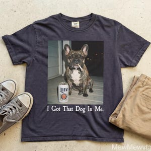 May include: Dark navy blue t-shirt featuring a graphic of a brindle French bulldog with a cigarette in its mouth, a can of Lite beer, and the text "I Got That Dog In Me." The shirt is paired with khaki pants and gray sneakers.