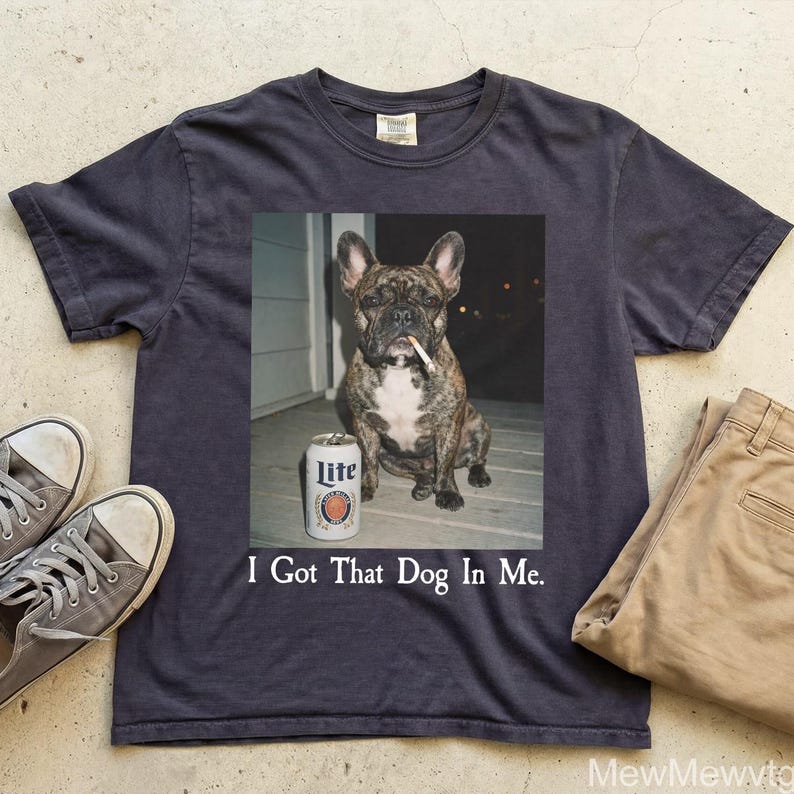 May include: Dark navy blue t-shirt featuring a graphic of a brindle French bulldog with a cigarette in its mouth, a can of Lite beer, and the text "I Got That Dog In Me." The shirt is paired with khaki pants and gray sneakers.