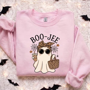 Pode incluir: Uma camisola de gola redonda rosa claro com um gráfico de um fantasma a usar um lenço com padrão de leopardo e óculos de sol redondos. O fantasma segura um café gelado e um saco com padrão de leopardo. O texto BOO-JEE está impresso acima.