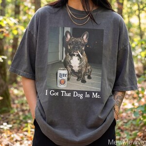 May include: Dark grey t-shirt featuring a graphic of a brindle French Bulldog with a cigarette in its mouth and a can of Lite beer. The text "I Got That Dog In Me" is printed below the image.