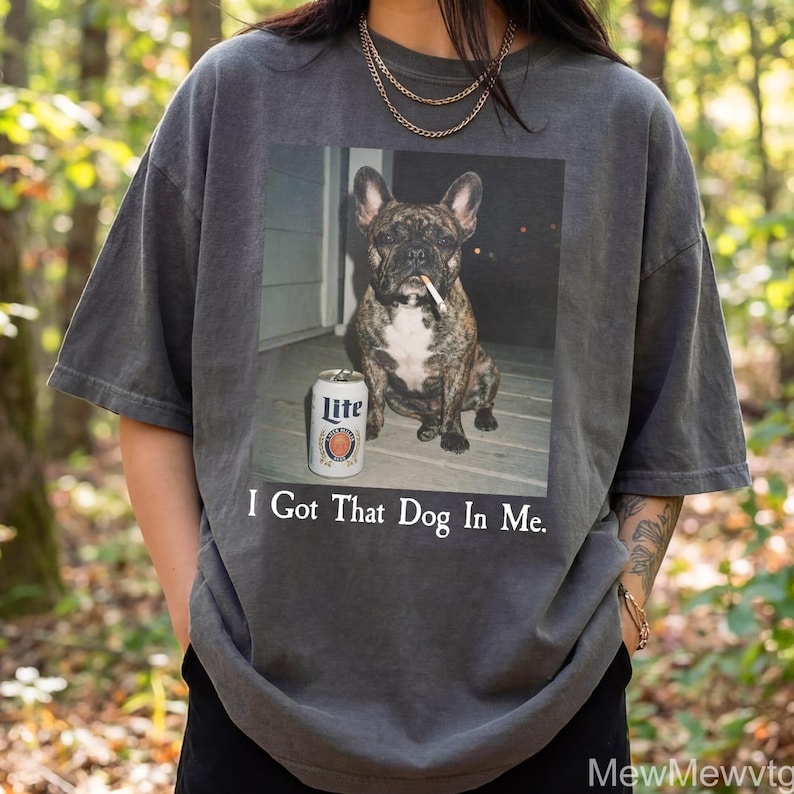 May include: Dark grey t-shirt featuring a graphic of a brindle French Bulldog with a cigarette in its mouth and a can of Lite beer. The text "I Got That Dog In Me" is printed below the image.