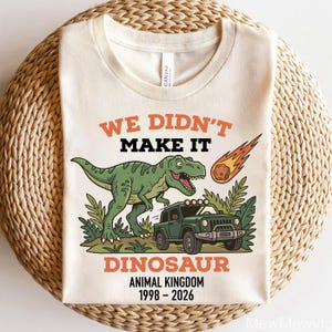 Może przedstawiać: Kremowy t-shirt z zielonym Tyrannosaurusem Rexem, zielonym jeepem i płonącym meteorem. Tekst brzmi "WE DIDN'T MAKE IT DINOSAUR ANIMAL KINGDOM 1998-2026" w kolorze pomarańczowym i czarnym.