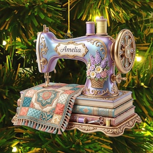 Pode incluir: Um ornamento decorativo em forma de máquina de costura lavanda com detalhes dourados, apresentando o nome Amelia, uma cesta de flores e uma peça de tecido acolchoado com borlas sobre uma pilha de três livros com pequenas ferramentas de costura.