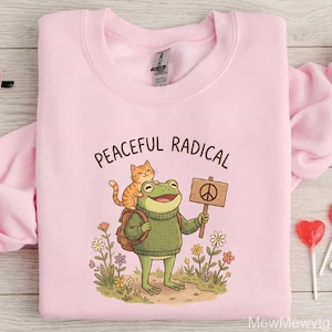 May include: A light pink sweatshirt with the words "PEACEFUL RADICAL" printed across the top. The graphic features a green frog wearing a sweater and backpack, holding a peace sign, with an orange cat on its head. The design is surrounded by flowers.