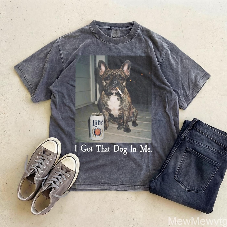May include: A gray t-shirt with a graphic of a French Bulldog with a cigarette and a can of Lite beer. The text "I Got That Dog In Me" is below the image. The shirt is paired with gray sneakers and dark wash jeans.