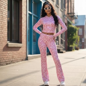 Puede incluir: Un conjunto de dos piezas con estampado de leopardo rosa, que incluye un top corto con capucha y pantalones acampanados. El conjunto se usa con zapatillas blancas y gafas de sol. El fondo presenta un edificio de ladrillo y una puerta azul.
