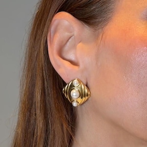 Könnte beinhalten: Ein goldfarbener Ohrring mit einem rautenförmigen, gerippten Metalldesign, das drei weiße, runde, abgestufte Perlen aufweist, die vertikal in der Mitte angeordnet sind. Der Ohrring hat eine polierte Oberfläche und einen Clip-Verschluss.