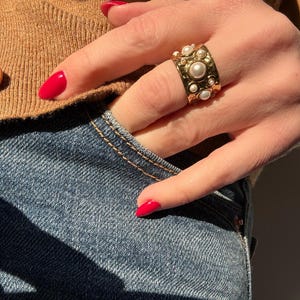 Könnte beinhalten: Ein goldfarbener Ring mit mehreren Kunstperlen wird an einem Finger getragen. Der Ring hat eine strukturierte Oberfläche und ein breites Band. Die Person trägt einen braunen Pullover und eine blaue Jeans. Die Nägel sind rot lackiert.