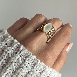 Könnte beinhalten: Ein goldfarbener Ring mit einem ovalen, perlmuttartigen Stein. Der Ring hat ein detailliertes, durchbrochenes Design um den Stein und das Band. Der Ring wird an einer Hand getragen, mit einem weißen Strickärmel im Hintergrund.