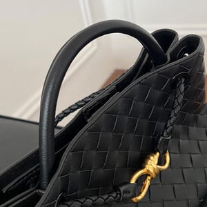 K&ouml;nnte beinhalten: Nahaufnahme einer schwarzen, gewebten Leder-Tote-Tasche mit gebogenem Griff und geflochtenen Schulterriemen. Die Tasche hat einen goldfarbenen Verschluss und ein strukturiertes Design. Das Bild zeigt die Textur und Verarbeitung der Handtasche.