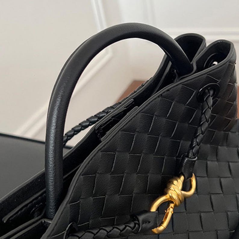 K&ouml;nnte beinhalten: Nahaufnahme einer schwarzen, gewebten Leder-Tote-Tasche mit gebogenem Griff und geflochtenen Schulterriemen. Die Tasche hat einen goldfarbenen Verschluss und ein strukturiertes Design. Das Bild zeigt die Textur und Verarbeitung der Handtasche.