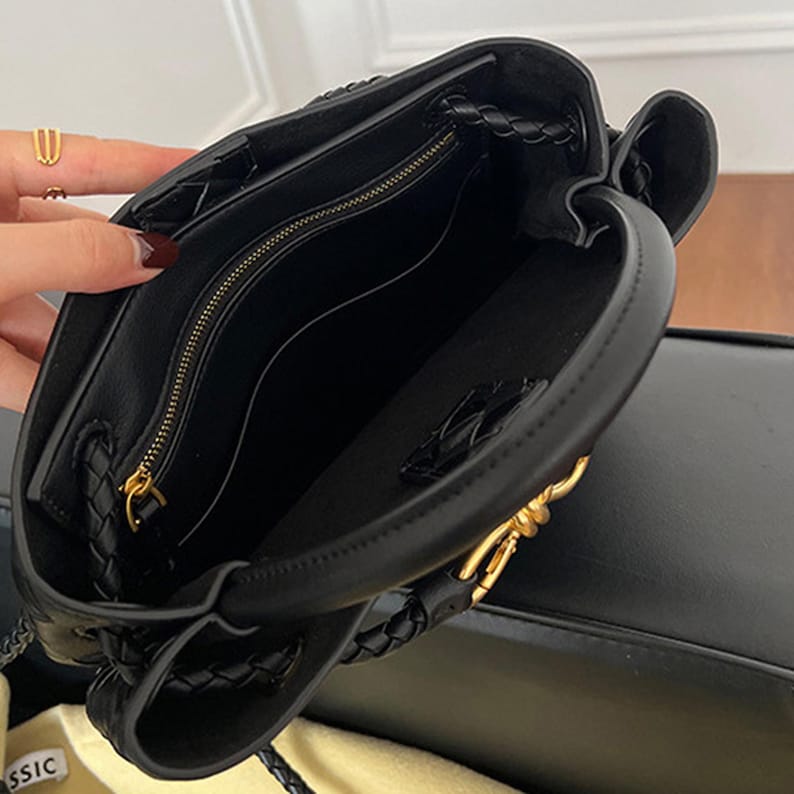 K&ouml;nnte beinhalten: Schwarze Lederhandtasche mit geflochtenem Griff und goldfarbener Hardware. Der Innenraum verf&uuml;gt &uuml;ber eine Rei&szlig;verschlusstasche. Die Tasche ist offen und zeigt das Innenfutter. Die Tasche steht auf einer schwarzen Oberfl&auml;che.