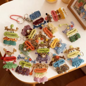 Puede incluir: Una colección de llaveros de ganchillo hechos a mano y coloridos en varios diseños. Cada llavero presenta una criatura única y caprichosa con rasgos expresivos y combinaciones de colores vibrantes, incluyendo verde, naranja y azul. Los llaveros están dispuestos sobre una superficie blanca.