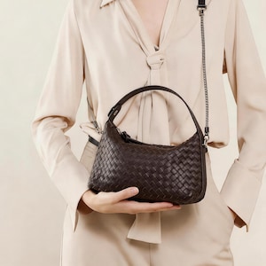 May include: A dark brown woven leather shoulder bag with a curved shape. The bag has a top handle and a silver chain strap. The bag is held by a person wearing a beige blouse and pants.