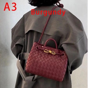 K&ouml;nnte beinhalten: Burgunderfarbene Handtasche mit gewebtem Design. Die Tasche hat einen oberen Griff und einen Schulterriemen. Ein goldfarbener Verschluss ziert die Vorderseite. Die Texte "A3" und "Burgundy" sind im Bild zu sehen.