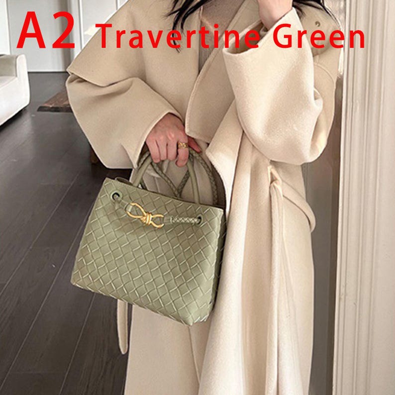 K&ouml;nnte beinhalten: Hellgr&uuml;ne, gewebte Handtasche mit goldfarbenem Verschluss. Die Tasche hat einen geflochtenen Griff und einen Kordelzugverschluss. Der Text "A2 Travertine Green" ist sichtbar. Eine Person in einem cremefarbenen Mantel h&auml;lt die Tasche.