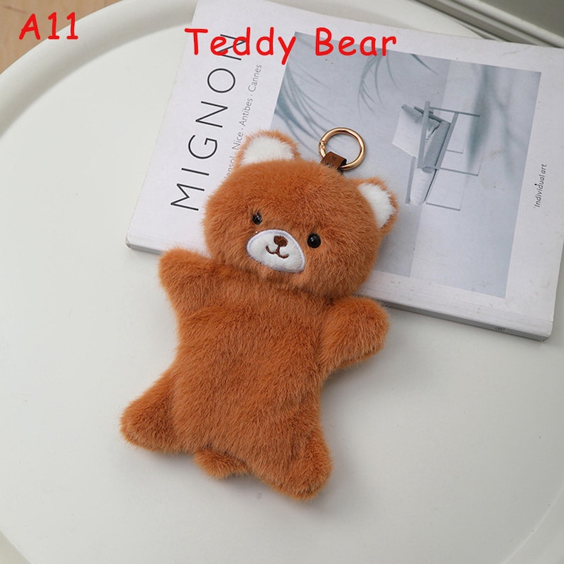 K&ouml;nnte beinhalten: Ein brauner Teddyb&auml;r-Schl&uuml;sselanh&auml;nger mit wei&szlig;en Akzenten an den Ohren. Der Pl&uuml;schb&auml;r hat ein aufgesticktes Gesicht und einen goldfarbenen Schl&uuml;sselring. Der Text "Teddy Bear" steht &uuml;ber dem B&auml;ren.