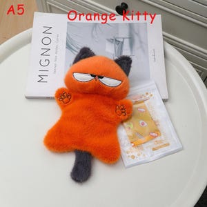 K&ouml;nnte beinhalten: Ein orangefarbenes Pl&uuml;schk&auml;tzchen mit grimmigem Gesicht, schwarzen Ohren und einem grauen Schwanz. Die Katze hat gestickte Augen und Pfoten. Der Text "Orange Kitty" steht &uuml;ber der Katze. Ein Buch und eine kleine Packung sind im Hintergrund.