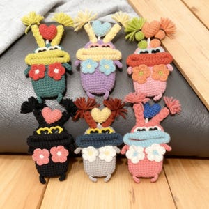 May include: A collection of six handmade crochet dolls in various colors. Each doll features a unique design with heart-shaped accents, flower details, and yarn hair. The dolls are arranged on a wooden surface and a gray cushion.