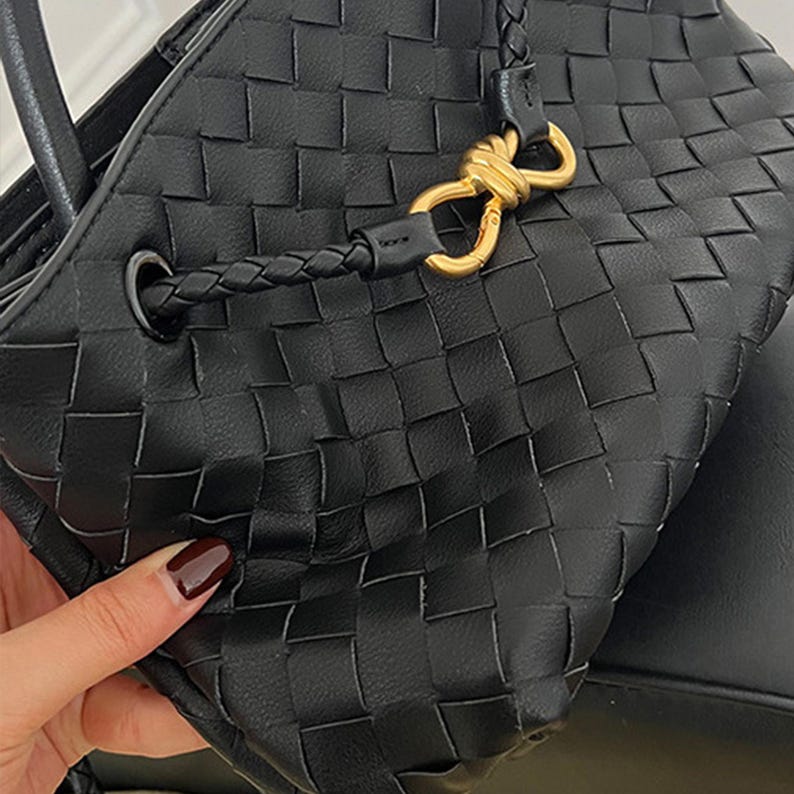 K&ouml;nnte beinhalten: Schwarze Handtasche aus geflochtenem Leder mit geflochtenem Griff und goldfarbener Hardware. Die Tasche hat ein einzigartiges Korbmuster und einen sicheren Verschluss. Das Design ist modern und stilvoll.