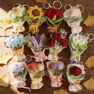 May include: A collection of handmade crochet flower keychains in various colors, including white, yellow, red, pink, blue, and purple. Each miniature bouquet is wrapped with a ribbon and features a gold-colored keychain ring.