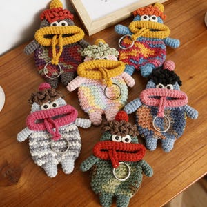 Puede incluir: Una colección de llaveros de ganchillo hechos a mano con forma de criaturas caprichosas. Cada llavero presenta una combinación de colores única, incluyendo arcoíris, azul y verde, con una boca de color contrastante y un anillo metálico para llaves.