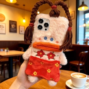 Puede incluir: Una funda de teléfono novedosa con forma de pato de dibujos animados. La funda rosa tiene trenzas marrones, un pico naranja y una falda roja con detalles de flores amarillas. La funda tiene una textura esponjosa.