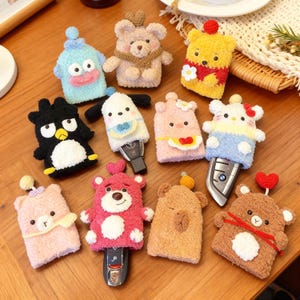 May include: A collection of plush key covers in various character designs, including bears, penguins, and other cartoon figures. The covers are in a variety of colors, such as pink, yellow, and black, and are designed to fit over car keys.