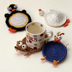 Peut inclure: Un mug en céramique blanc à pois colorés avec le texte MRS, entouré de quatre dessous de verre crochetés en forme d'animaux, incluant un pingouin noir, un oiseau blanc, une créature brune aux grands yeux et un oiseau bleu aux pattes orange.