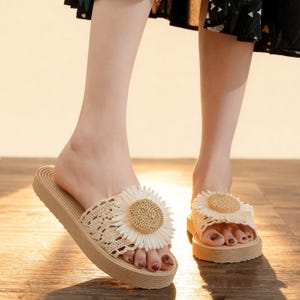 May include: Tan platform sandals with a crochet strap and a large, cream-colored daisy embellishment. The sandals have a thick sole and are designed for casual wear. The daisy has a woven center.