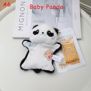 K&ouml;nnte beinhalten: Ein Pl&uuml;sch-Panda-Schl&uuml;sselanh&auml;nger mit wei&szlig;em K&ouml;rper und schwarzen Akzenten. Der Panda hat ein niedliches Gesicht und tr&auml;gt ein wei&szlig;es Outfit mit einem Cartoon-Motiv. Der Schl&uuml;sselanh&auml;nger ist an einem goldfarbenen Ring befestigt. Der Text "Baby Panda" ist sichtbar.
