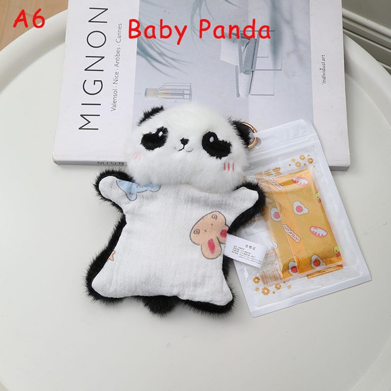 K&ouml;nnte beinhalten: Ein Pl&uuml;sch-Panda-Schl&uuml;sselanh&auml;nger mit wei&szlig;em K&ouml;rper und schwarzen Akzenten. Der Panda hat ein niedliches Gesicht und tr&auml;gt ein wei&szlig;es Outfit mit einem Cartoon-Motiv. Der Schl&uuml;sselanh&auml;nger ist an einem goldfarbenen Ring befestigt. Der Text "Baby Panda" ist sichtbar.