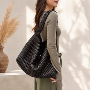 Puede incluir: Un bolso de hombro grande de cuero negro tejido con una correa trenzada. El bolso se lleva sobre el hombro y tiene un cierre de botón. El bolso es de color oscuro y parece estar vacío. El bolso es un accesorio elegante.