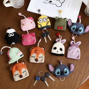 May include: A collection of colorful, crocheted keychains in various character designs, including a Minion, Hello Kitty, a frog, and a panda. Each keychain has a metal ring for keys and a small loop at the top.