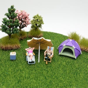 Pode incluir: Um diorama em miniatura com duas pequenas figuras relaxando sob um guarda-sol bege em uma superfície de grama verde. Uma figura veste um top rosa e shorts brancos em uma espreguiçadeira, a outra uma camisa bege perto de uma barraca roxa.