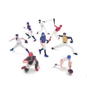 May include: A collection of small baseball player figurines in action poses. The figures wear team uniforms in colours like red, blue, white, and yellow. The scene includes players pitching, batting, and fielding on a white background.