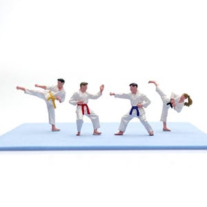 May include: Miniature figures in white karate uniforms with colored belts demonstrate martial arts poses on a light blue mat. The figures include a yellow, red, blue, and black belt. The background is white.