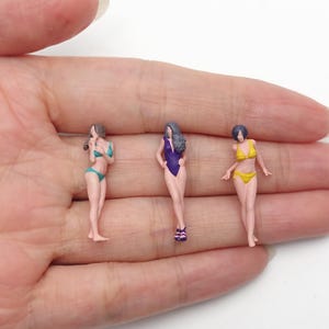 Op de afbeelding: Drie miniatuurfiguurtjes van mensen die op een rij staan. Het eerste figuurtje draagt een tweekleurig blauwgroen zwempak, het tweede een paars badpak met paarse sandalen en het derde een geel tweekleurig zwempak.