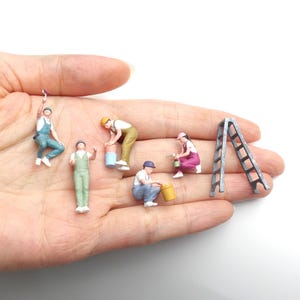 Puede incluir: Un conjunto de cinco figuras en miniatura que representan trabajadores de la construcción en varias posturas, incluyendo figuras con rodillos de pintura, cubos y una escalera. Las figuras llevan monos y gorros coloridos, a pequeña escala.