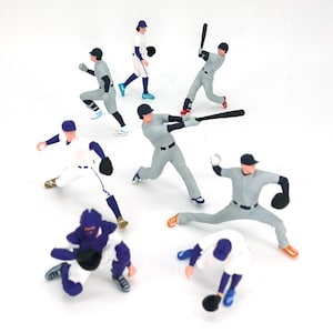 Peut inclure: Plusieurs figurines miniatures de joueurs de baseball en pleine action. Les figurines portent des uniformes de baseball, avec un mélange de couleurs grises, blanches et bleues. Certains battent, lancent, courent et défendent.