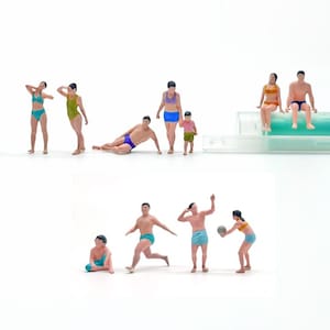 May include: Miniature figurines of people in swimwear, engaged in various beach activities. The scene includes figures sunbathing, playing with a ball, and sitting, all set against a white background. The figurines are wearing swimsuits in various colors.